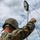 Marines with 3d Supply Battalion demonstrate their field communication capabilities Two military personnel in MARPAT digital camouflage uniforms are setting up what appears to be a satellite or communications dish antenna on an outdoor paved surface. The foreground soldier, whose helmet tape reads 'AGUALDE' (partially visible), is holding guide wires to stabilize the antenna mast. The scene takes place under a partly cloudy sky with vegetation visible in the background.