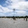 Marines with 3d Supply Battalion demonstrate their field communication capabilities Military personnel are setting up a field communications site on a gravel surface, featuring a large parabolic dish antenna mounted on a tall mast secured with guy wires. Several soldiers in camouflage uniforms are working around the equipment, which includes military vehicles, a tent structure, and additional antenna masts. The setting appears to be an outdoor training or operational area bordered by dense vegetation under a partly cloudy sky.