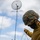 Marines with 3d Supply Battalion demonstrate their field communication capabilities A U.S. Marine in the foreground wearing MARPAT camouflage helmet, black face covering, and olive drab fleece is handling guide wires or antenna cables. In the background, several other Marines are working together to erect what appears to be a tall antenna mast topped with a parabolic dish or grid antenna. The scene takes place outdoors on a flat surface under a partly cloudy sky.