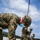 Marines with 3d Supply Battalion demonstrate their field communication capabilities A U.S. Marine in MARPAT digital camouflage uniform and blue surgical face mask is bent over handling a green rope or cord on what appears to be a gravel or tarmac surface. In the background, a group of similarly uniformed personnel are gathered and working with lines or rigging equipment. The setting appears to be an outdoor training or operational exercise, likely involving aerial rigging or antenna setup.