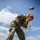 Marines with 3d Supply Battalion demonstrate their field communication capabilities A female service member wearing USMC MARPAT digital camouflage uniform and tan combat boots is driving a metal stake into a gravel surface using a yellow-handled sledgehammer. The low-angle photograph captures her in a wide stance, leaning forward with focused effort. The setting appears to be an outdoor military work site under a partly cloudy blue sky.