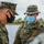 Marines with 3d Supply Battalion demonstrate their field communication capabilities Two U.S. Marines in MARPAT digital camouflage uniforms are engaged in conversation in an outdoor, wetland or coastal environment. Both personnel are wearing face masks, consistent with COVID-19 era protocols. The Marine on the right is identified by a name tape reading 'GREER' and wears a boonie cover, while the Marine on the left wears a patrol cap.
