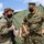 Marines with 3d Supply Battalion demonstrate their field communication capabilities The image shows U.S. Marines in MARPAT digital camouflage uniforms wearing black face masks, gathered near a large military field tent. Two primary figures are engaged in conversation, with one holding documents and a rifle, while others are visible in the background. The setting appears to be an outdoor military field exercise or operational briefing.