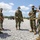 Marines with 3d Supply Battalion demonstrate their field communication capabilities Five military personnel in MARPAT woodland camouflage uniforms are gathered on a gravel surface, appearing to be in a briefing or debriefing situation. All individuals are wearing black face masks, consistent with COVID-19 era protocols. Military vehicles are visible in the background, suggesting a field exercise or operational environment.
