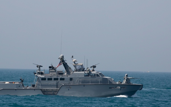 The image shows two gray military patrol boats underway on open blue water, appearing to be U.S. Navy or Coast Guard vessels based on the visible American flag. The boats are equipped with multiple weapon mounts, sensor equipment, and communications antennas, consistent with riverine or coastal patrol craft. The vessels are traveling in close formation, generating visible bow wakes.