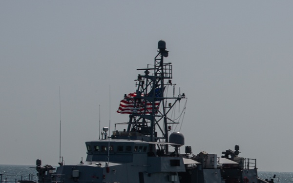 A U.S. Navy warship bearing hull number '3' is underway on open, choppy seas. The vessel is a gray-painted patrol coastal or gunboat class ship flying the American flag from its mast. The image was taken from another vessel at close range under overcast conditions.