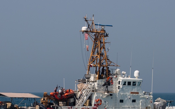 A U.S. Coast Guard patrol vessel is operating at sea alongside a smaller vessel named 'ADAK'. The Coast Guard cutter, bearing hull number 1333, appears to be conducting an at-sea boarding or interdiction operation, with personnel and an orange inflatable boat visible on deck. The scene takes place in open blue water under a hazy sky.