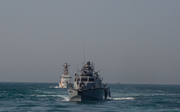 The image shows two military/patrol vessels underway on open ocean waters under a hazy sky. The foreground vessel is a dark gray armed patrol boat equipped with weapons systems and sensor masts, while a white-hulled cutter-type vessel follows in the background. The vessels appear to be conducting a joint patrol or transit operation.