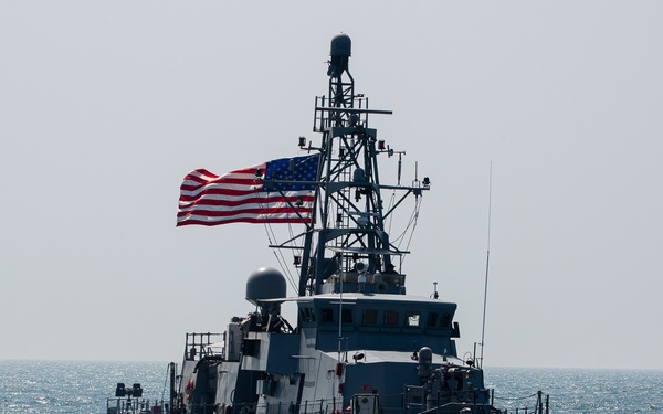 A U.S. Navy warship bearing hull number '3' is underway on open ocean waters. The vessel is flying a large American flag from its mast and appears to be a patrol coastal or similar small combatant vessel painted in standard naval gray. The ship is moving at moderate speed, creating a visible bow wake and stern wake.