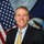 A formal portrait photograph of a middle-aged man in professional civilian attire, posed in front of a United States flag and what appears to be a U.S. Navy flag. The setting and composition are consistent with an official government or military civilian leadership portrait. The subject is smiling and dressed in a dark suit with a light blue shirt and gold patterned tie.