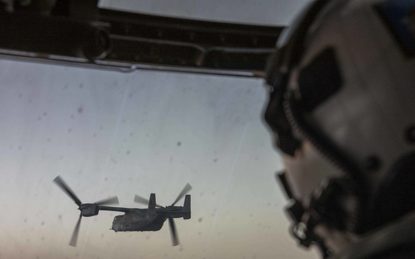 A military pilot or crew member is seated in the cockpit of an aircraft, observing a V-22 Osprey tiltrotor aircraft flying in formation ahead. The image is taken from inside the cockpit looking forward through the windscreen, with the Osprey visible in the near distance against a hazy twilight sky. The cockpit displays multiple illuminated multifunction displays and avionics panels.