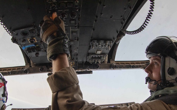 Two military aircrew members are photographed inside a helicopter cockpit during flight, captured with a fisheye lens. The right-seat pilot is reaching up to manipulate overhead panel controls while wearing full flight gear including helmet and communications equipment. Multiple green-lit avionics displays are visible on the instrument panel.