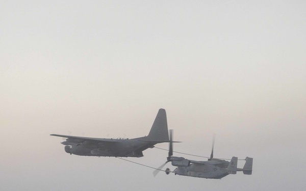 The image shows two military aircraft conducting an aerial refueling operation in hazy, overcast conditions. A large four-engine turboprop tanker aircraft, consistent with a KC-130 variant, is connected via a refueling hose and drogue to a tiltrotor aircraft consistent with a V-22 Osprey. The scene appears to be captured at dusk or dawn, with muted pinkish-gray tones in the sky.
