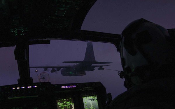 A military aircrew member is visible in silhouette in a darkened cockpit during what appears to be an aerial refueling operation at dusk or dawn. Multiple illuminated multifunction displays and avionics screens cast green light across the instrument panel. Through the windscreen, a large tanker aircraft — consistent with a KC-130 or similar platform — is visible with a refueling drogue extended toward the receiving aircraft.