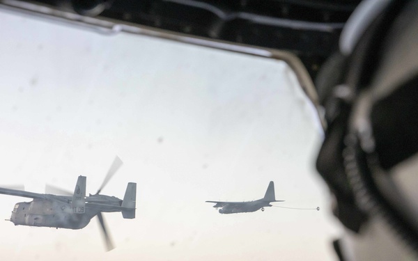 The image is taken from inside a military aircraft cockpit, showing a view through the windshield of two aircraft in flight. A V-22 Osprey tiltrotor aircraft is visible in the foreground with its rotors spinning, while a C-130 tanker aircraft is seen in the background with a refueling drogue extended. A helmeted crew member is partially visible on the right side of the frame.