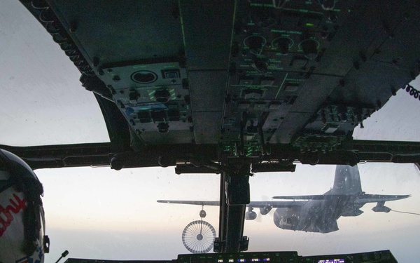 Two military pilots are seated in the cockpit of a military aircraft, viewed from behind, conducting an aerial refueling operation. A tanker aircraft is visible through the windscreen with a deployed refueling drogue basket extended toward the receiving aircraft. The cockpit displays multiple illuminated multifunction displays (MFDs) with green and blue navigation and flight data.