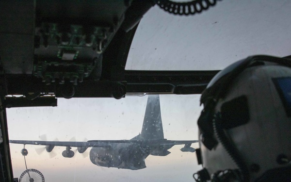 A military pilot in full flight gear is visible from behind in the cockpit of a rotary-wing aircraft, approaching a fixed-wing tanker aircraft (consistent with a C-130 variant) for aerial refueling. A refueling drogue/basket is visible extended from the tanker on a hose. The cockpit displays multiple green-illuminated multifunction displays and instrument panels, suggesting a night or low-light operation environment.