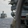 The image shows a view from the deck of a naval warship looking toward a large auxiliary or replenishment vessel in the open ocean. The foreground reveals military equipment including sensors, radar systems, and a life ring mounted on the warship's superstructure. A crew member is partially visible near the equipment on the right side of the frame.