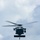 A military helicopter is photographed head-on in flight against an overcast sky. The aircraft appears to be a Sikorsky MH-60 Seahawk or similar variant, equipped with external stores or sensor pods mounted beneath the fuselage. The rotor blades show motion blur, indicating active flight.