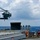 A military helicopter, consistent with an MH-60 Seahawk, is hovering or approaching to land on the deck of a naval warship at sea. The ship's deck features various equipment including a life ring, cable reels, and what appears to be a missile launcher system. The scene takes place in open ocean under a partly cloudy sky.