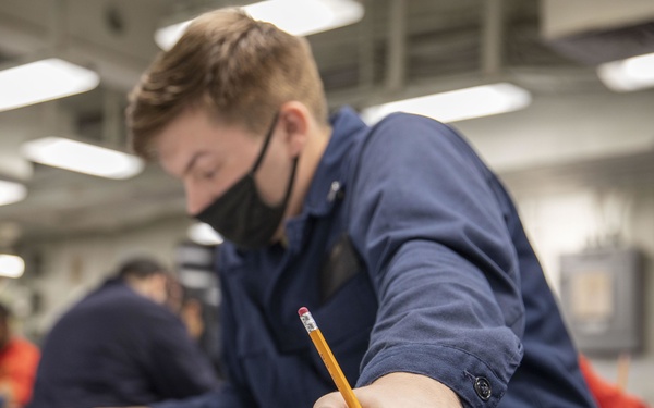 A young person wearing a black face mask and navy blue uniform is seated at a green-topped desk, appearing to take a written exam or complete paperwork. They are holding a yellow pencil and have additional pencils on the desk surface. The setting appears to be a military or institutional testing environment with multiple individuals visible in the background.