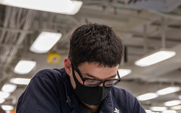 A person wearing a dark navy uniform, black face mask, and glasses is seated at a table writing with a pencil on documents or a workbook. The setting appears to be a large indoor facility, likely a military vessel or institutional space, with fluorescent lighting and other individuals visible in the background. The scene suggests a formal testing, training, or administrative activity.