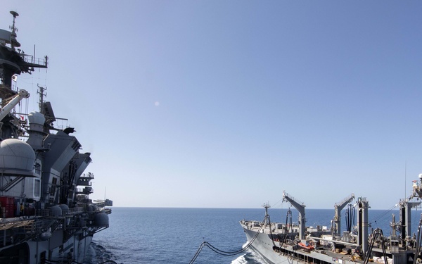 Two naval vessels are conducting an underway replenishment (UNREP) operation at sea, with multiple fuel/transfer hoses connected between them. The ship on the left appears to be a large amphibious assault ship, while the vessel on the right is a replenishment oiler or combat stores ship. Both ships are underway in calm, blue ocean waters under clear skies.