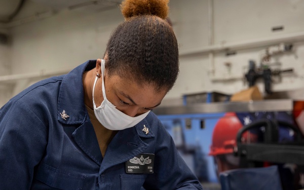 A U.S. Navy sailor in blue working uniform is seated at a workbench, writing or reviewing documents using a ruler and pencil. The sailor is wearing a white face mask and has visible rank insignia and a name tag reading 'RONNESHA' on their uniform. The setting appears to be an interior workspace or workshop aboard a naval vessel.