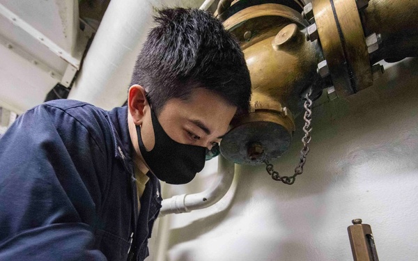 A person wearing a dark blue uniform and black face mask is performing maintenance or inspection work on brass/bronze valves and piping systems in what appears to be a naval vessel's interior compartment. The individual has visible tattoos on their right forearm and is closely examining or adjusting a brass valve assembly. The setting is consistent with a ship's engineering or mechanical space.