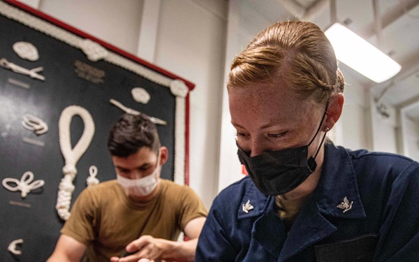 Two military personnel, likely U.S. Navy sailors, are working in what appears to be a shipboard compartment or training room. A female sailor in navy blue coveralls and a black face mask is inspecting or tagging a white container, while a male sailor in a tan t-shirt and white face mask works with orange safety equipment in the background. A knot-tying reference board is mounted on the wall behind them.