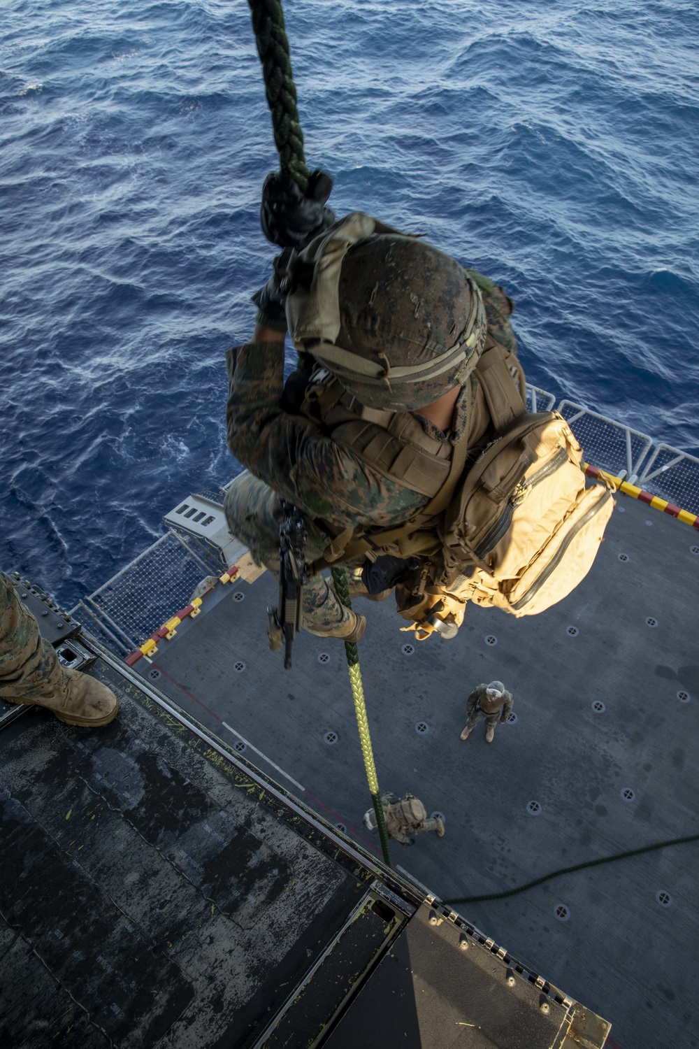 A military service member in full combat gear is rappelling down a rope from an elevated position onto what appears to be a ship's flight deck or platform over open ocean. The perspective is a top-down aerial view, likely taken from a helicopter, showing the soldier mid-descent with the blue ocean visible in the background. At least two additional personnel are visible on the deck below.