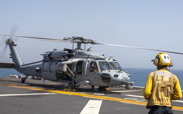 A military helicopter, marked with the number 52, is positioned on the flight deck of a naval vessel at sea. Two flight deck crew members are visible — one in a yellow jersey and yellow helmet, the other in a green jersey and green helmet — monitoring the helicopter. The scene appears to be a pre-launch or post-landing operation under clear, hazy sky conditions.