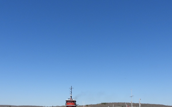 A classic red-and-black tugboat operates on a calm river or harbor alongside a small workboat and a large floating platform or barge with personnel on deck. The scene appears to depict a marine operation, possibly a vessel delivery, docking maneuver, or infrastructure installation. A wooded hillside and scattered buildings are visible in the background under a clear blue sky.