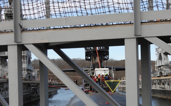 The image shows a submarine in a naval shipyard or dry dock facility, viewed through a steel structural framework. Workers in safety gear are visible on and around the submarine, which appears to be undergoing maintenance or overhaul operations. Crane equipment and industrial infrastructure are present on both sides of the waterway.