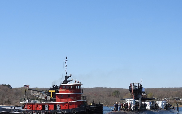 A red commercial tugboat with a yellow 'T' marking on its stack is assisting a dark-hulled military submarine in a river or coastal waterway. Personnel are visible on the submarine's deck near the sail (conning tower), suggesting an active docking, undocking, or transit operation. The scene is set against a backdrop of bare deciduous trees and a clear blue sky, indicating early spring conditions.
