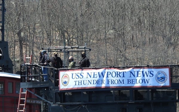 The image shows a naval ceremony or launch event for the USS Newport News submarine, with a large banner reading 'USS Newport News - Thunder From Below' displayed on what appears to be a floating dry dock or support structure. Several personnel are visible on an elevated platform, and a red tugboat identified as 'Paul A. Wronowski' is positioned alongside. The setting appears to be a shipyard or naval facility, likely during late winter or early spring based on the bare deciduous trees in the background.