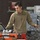 A young man wearing an olive/tan military-style quarter-zip fleece is working in what appears to be a military maintenance or technical facility. He is operating equipment at a workstation that includes a ruggedized laptop and orange-colored test equipment. The background shows storage cabinets, warning signs, and various technical equipment typical of a military hangar or maintenance bay.