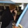A technician or mechanic is performing maintenance work on an aircraft, inspecting an internal compartment using a handheld flashlight or inspection light. The worker is dressed in a black long-sleeve shirt and an olive/mustard beanie hat, leaning into an open access panel on the underside or fuselage of the aircraft. Various wiring, ducting, and structural components are visible inside the open bay.
