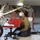 A technician wearing a surgical mask, head-mounted light, and blue gloves is performing maintenance on the undercarriage of an aircraft, inspecting the landing gear assembly from a kneeling position on the hangar floor. The worker is using a Snap-on mechanic's stool nearby and appears to be conducting a close visual inspection of the wheel well and strut components. The setting is an indoor aircraft hangar with the aircraft's nose gear area clearly visible.