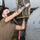 A male aircraft maintenance technician wearing a black face mask and olive/tan t-shirt is performing maintenance work on a military aircraft in what appears to be a hangar. He has visible tattoos on his right forearm and is reaching into an open access panel on the underside or side of the aircraft. The aircraft's gray fuselage, riveted panels, and engine components are clearly visible.