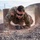 A U.S. Army soldier in full OCP (Operational Camouflage Pattern) uniform is low-crawling under a barbed wire obstacle on a gravel and dirt surface, kicking up dust as they move. The setting appears to be a military training course in an arid, desert-like environment. The soldier is wearing tactical gloves, ballistic eyewear, and displays unit patches including a U.S. flag patch on the right shoulder.