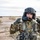A U.S. Army soldier in Operational Camouflage Pattern (OCP) uniform is adjusting a flight helmet with a microphone boom, suggesting aviation crew duties. The soldier displays a Sergeant rank insignia on the chest and an American flag patch on the right shoulder. In the background, a military helicopter (likely a UH-60 Black Hawk) is visible on an arid, desert-like terrain.