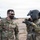 Two U.S. Army soldiers are engaged in conversation in an arid, desert-like outdoor environment. The soldier on the left, identified by name tape as 'Moore,' wears standard OCP camouflage with a black face mask and sunglasses, while the soldier on the right wears a flight helmet with a visor and appears to be a helicopter crew member. A military helicopter, likely a UH-60 Black Hawk, is visible in the background.