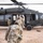 Military personnel in camouflage uniforms are approaching and boarding a UH-60 Black Hawk helicopter on an arid, desert terrain. The helicopter is on the ground with its side door open, and at least one crew member is visible in the cockpit. The scene appears to depict a troop loading or insertion operation in a dry, open landscape.