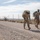 Two U.S. Army soldiers in MultiCam/OCP camouflage uniforms conduct a ruck march along a gravel patrol road adjacent to a border fence in an arid desert environment. Both soldiers carry heavy rucksacks and wear face masks, suggesting this was taken during the COVID-19 pandemic period. Additional soldiers are visible in the background performing the same activity along the fence line.
