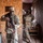 Two U.S. Army soldiers in MultiCam/OCP camouflage uniforms are conducting what appears to be a military training exercise inside a mock building structure made of OSB plywood and shipping containers. The foreground soldier is holding an M4-style rifle with a red training/safety device attached, indicating a simulated training environment. The second soldier is moving dynamically through a doorway into an adjacent corridor area.