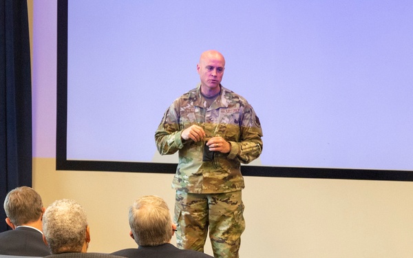A military service member in camouflage uniform is presenting or briefing a small group of civilians in an auditorium or briefing room. The audience, seated in the front rows of an otherwise largely empty venue, faces the presenter who stands in front of a large projection screen. The setting appears to be a formal military or government facility.