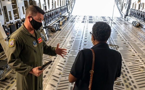 A military aircrew member in an olive green flight suit is conducting what appears to be a briefing or tour inside the cargo hold of a large military transport aircraft, likely a C-17 Globemaster III. He is speaking to a civilian visitor who is facing away from the camera and carrying a brown handbag. The interior of the aircraft cargo bay is clearly visible, showing the characteristic roller-track flooring and tie-down systems.