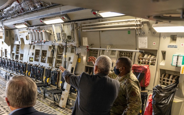 The image shows the interior of a large military cargo aircraft, likely a C-17 Globemaster III, where a military service member in camouflage uniform is briefing or explaining aircraft systems to two civilians in suits. The group is standing near the forward starboard wall of the cargo bay, which is lined with equipment panels, tie-down systems, and medical cross markings. The aircraft interior is well-lit and shows typical military transport configuration.