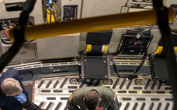 An overhead/bird's-eye view looking down into the cargo hold or interior of a large military or transport aircraft. Two individuals are visible on the aircraft floor — one in civilian attire and one in a military flight suit — appearing to conduct an inspection or briefing. The interior shows typical large aircraft features including tie-down tracks, an emergency exit sign, a fold-down crew seat, and safety equipment.
